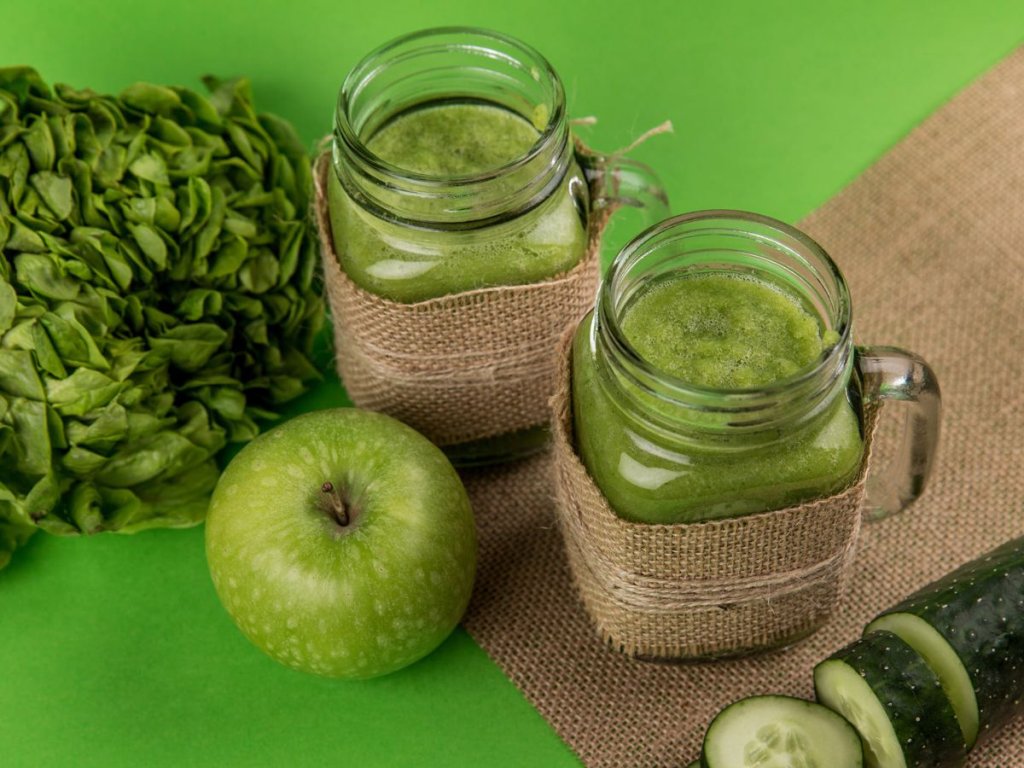 Ξύλινος πάγκος με πράσινο smoothie και δίπλα ένα αγγούρι, ένα πράσινο μήλο και ένα φύλλο καλέ
