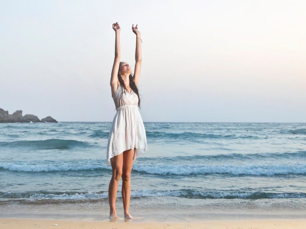 Γυναίκα σε παραλία με φόντο τη θάλασσα με λευκό φόρεμα και τα χέρια ψηλά