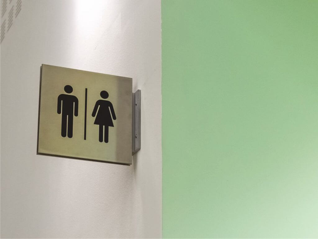 Τοίχος σε λευκό και πράσινο χρώμα με πινακίδα τουαλέτας που απεικονίζει με σκίτσο μια γυναικεία και μια ανδρική φιγούρα