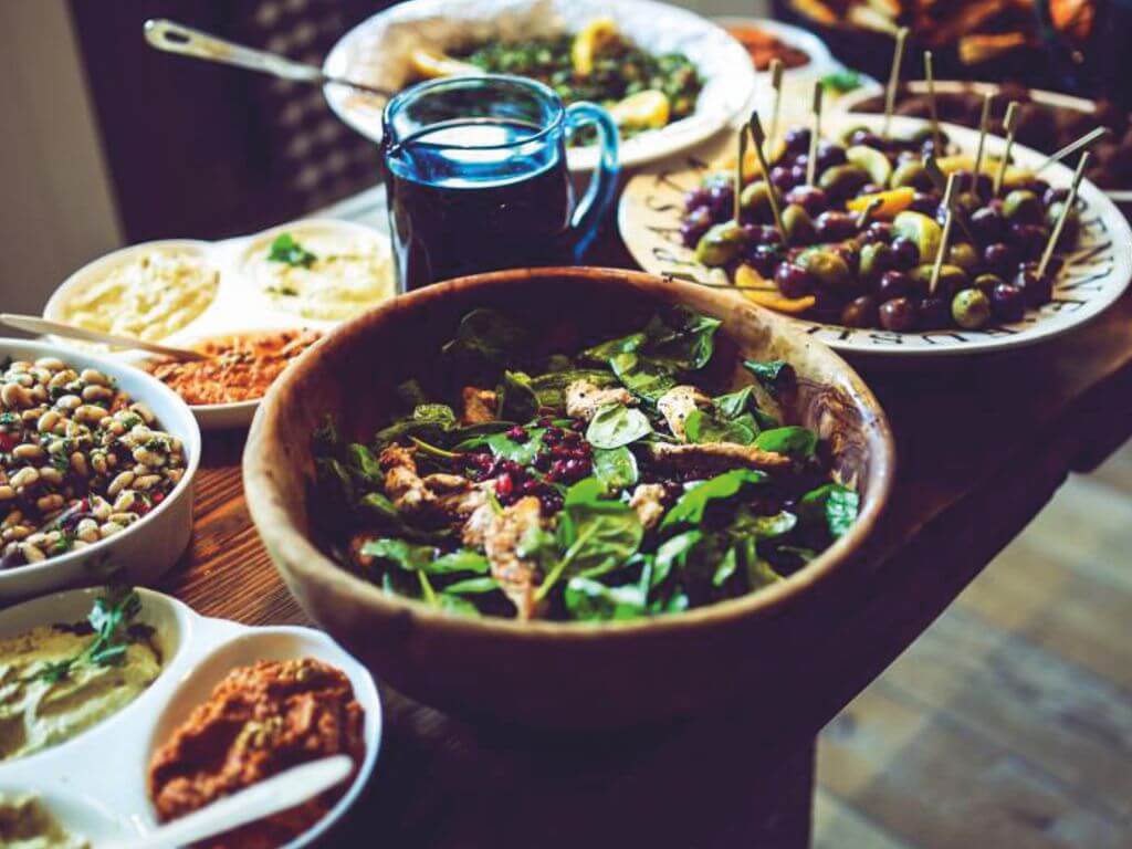Τραπέζι με διάφορα πιάτα, στο κέντρο υπάρχει μία πράσινη σαλάτα, δίπλα ένα πιάτο με ελιές, πίσω κι άλλες σαλάτες
