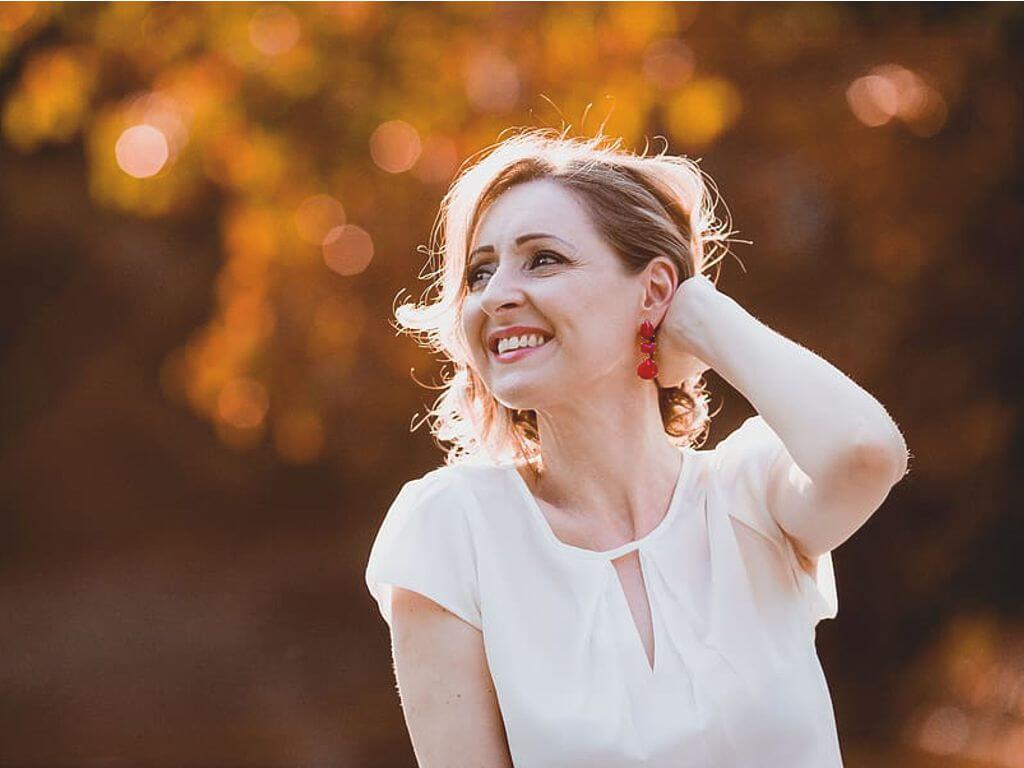 Ώριμη γυναίκα με λευκή μπλούζα φοράει κόκκινο κρεμαστό σκουλαρίκι, αγγίζει τα μαλλιά της και χαμογελάει