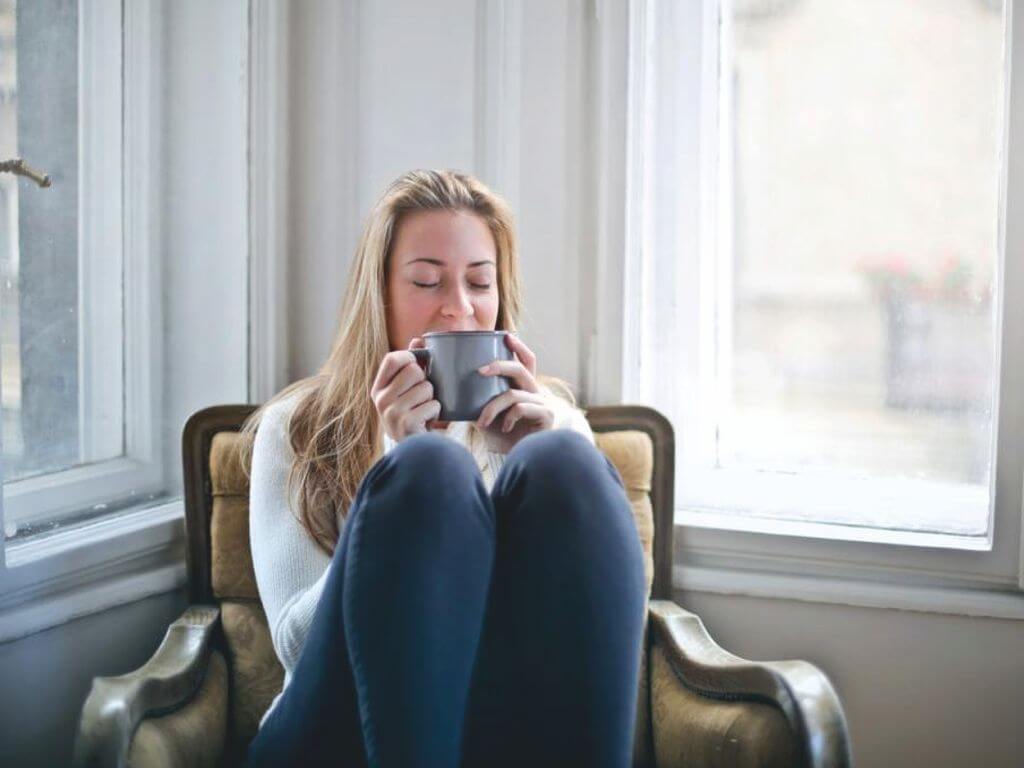 Ξανθιά γυναίκα με χαλαρή διάθεση και κλειστά μάτια καθισμένη σε μία πολυθρόνα κρατώντας μία γκρι κούπα