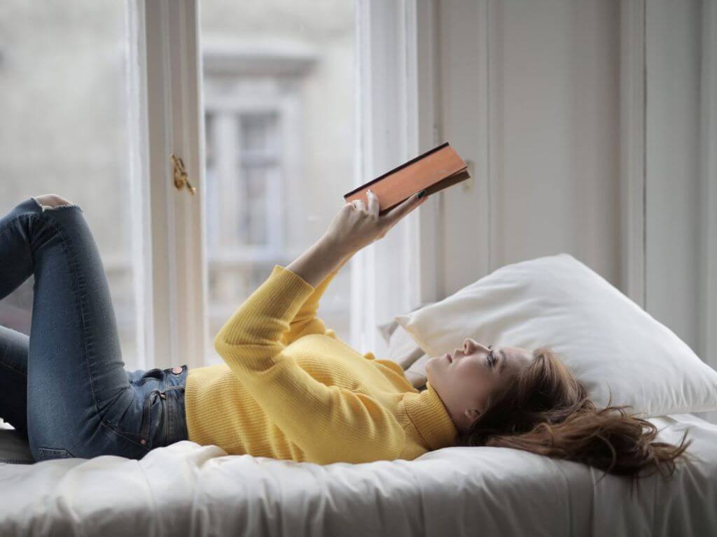Γυναίκα που διαβάζει ένα βιβλίο ξαπλωμένη στο κρεβάτι δίπλα σε ένα παράθυρο
