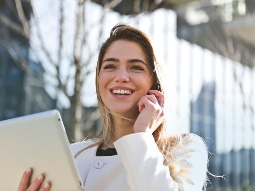 Χαμογελαστή ξανθιά γυναίκα μιλάει στο κινητό κρατώντας ένα tablet στο χέρι