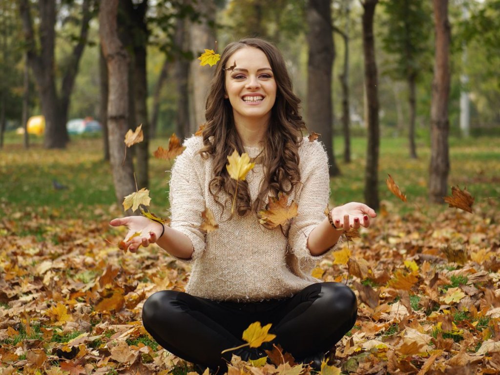Χαμογελαστή γυναίκα καθισμένη σε ένα πάρκο με ανοιχτά τα χέρια της και φύλλα που πέφτουν