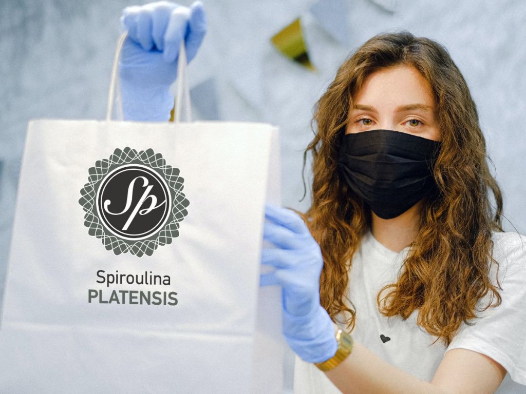 Γυναίκα με μαύρη ιατρική μάσκα στο πρόσωπο και μοβ γάντια μιας χρήσης κρατάει μία χάρτινη σακούλα αγορών με το λογότυπο Spiroulina PLATENSIS.