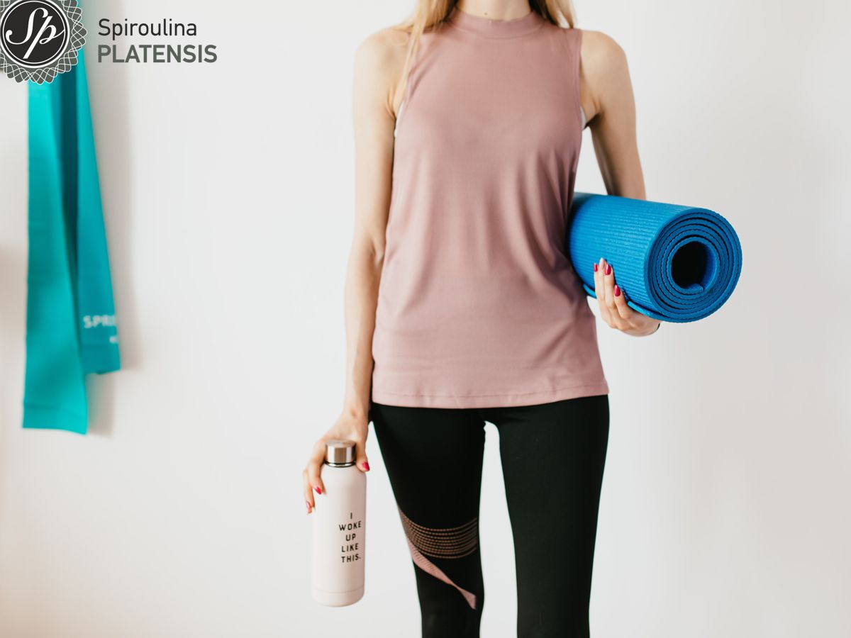 Νεαρή γυναίκα με αθλητικά ρούχα που κρατάει μπλε στρώμα γυμναστικής και ένα μπουκάλι