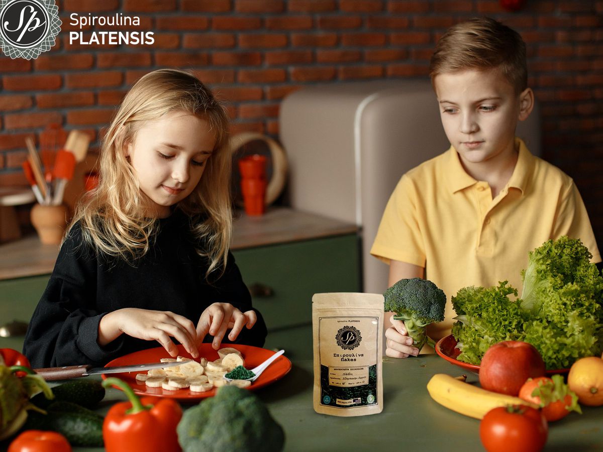 Δύο παιδιά, ένα αγόρι κι ένα κορίτσι που κάθονται πίσω από ένα τραπέζι που περιέχει πολλά φρούτα & λαχανικά & Σπιρουλίνα flakes