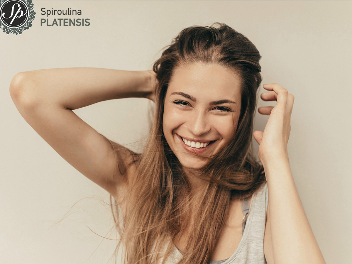 Χαμογελαστή γυναίκα με μακριά μαλλιά σε φωτεινό φόντο, με λογότυπο Spiroulina PLATENSIS, για άρθρο σχετικά με τη σπιρουλίνα και τη γυναίκα.