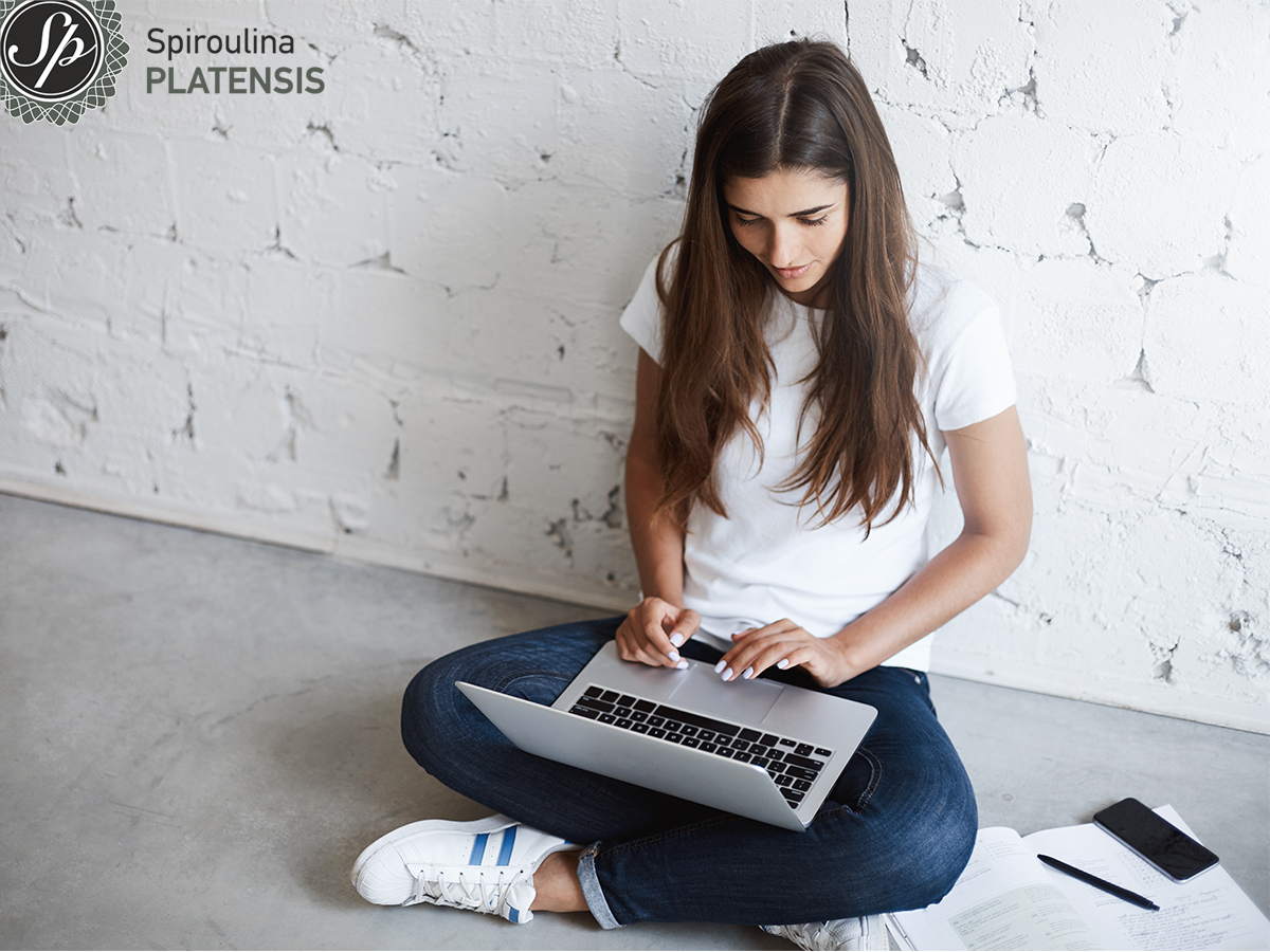 Νεαρή κοπέλα που κάθεται οκλαδόν με τον υπολογιστή στα πόδια που διαβάζει