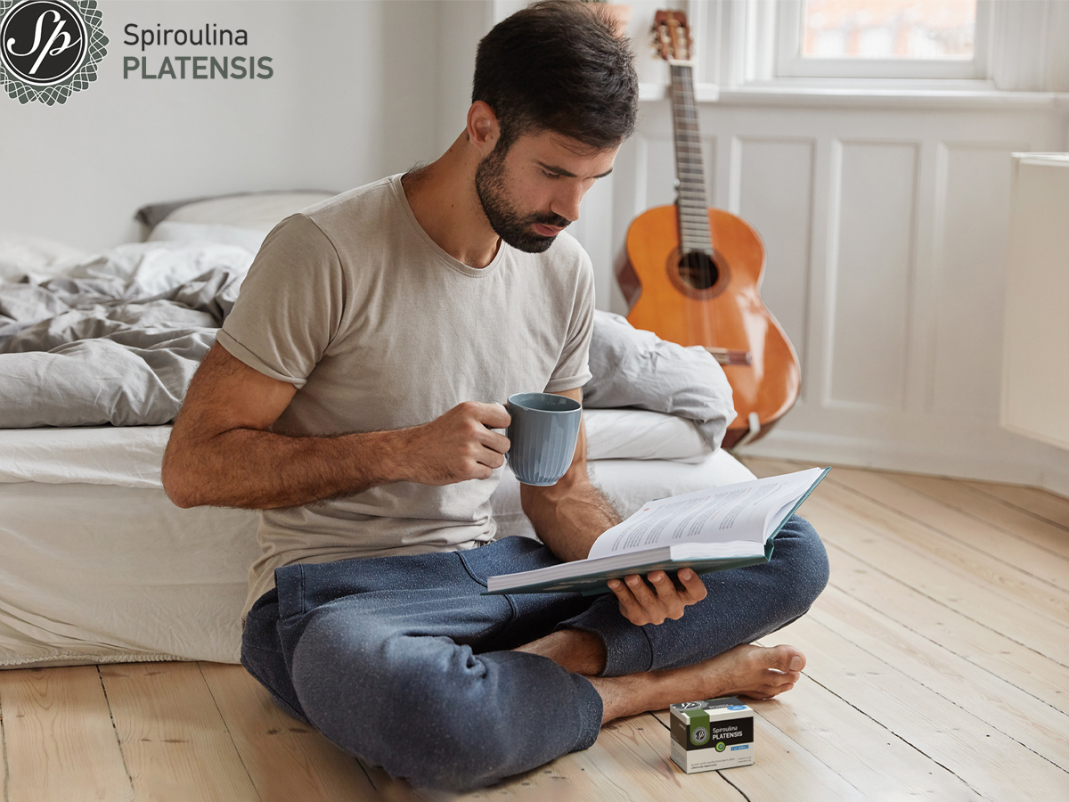 Άνδρας στο πάτωμα με κούπα και βιβλίο, δίπλα συσκευασία Spiroulina PLATENSIS
