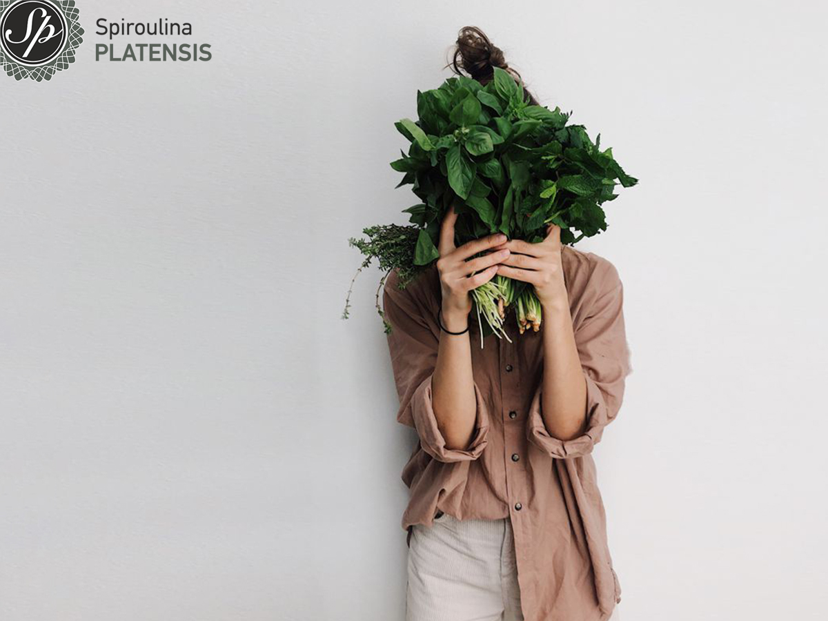 Νεαρή κοπέλα που στέκεται μπροστά από λευκό φόντο κρατόντας ένα μάτσο πράσινα λαχανικά κρύβοντας το πρόσωπό της