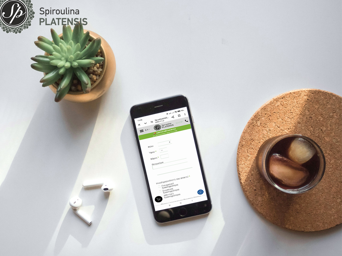Κινητό πάνω σε γραφείο με το Ερωτηματολόγιο Υγείας της Spiroulina PLATENSIS στην οθόνη, δίπλα σε ακουστικά, φυτό και ποτήρι με πάγο.