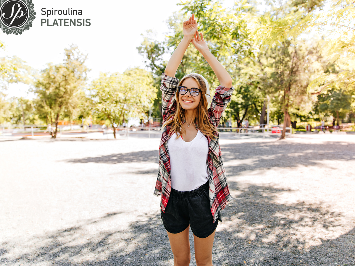 Νεαρή γυναίκα με μαύρο σορτς, λευκό tshirt, καρό πουκάμισο και γυαλιά σε εξωτερικό χώρο χαμογελάει με τα χέρια ψηλά