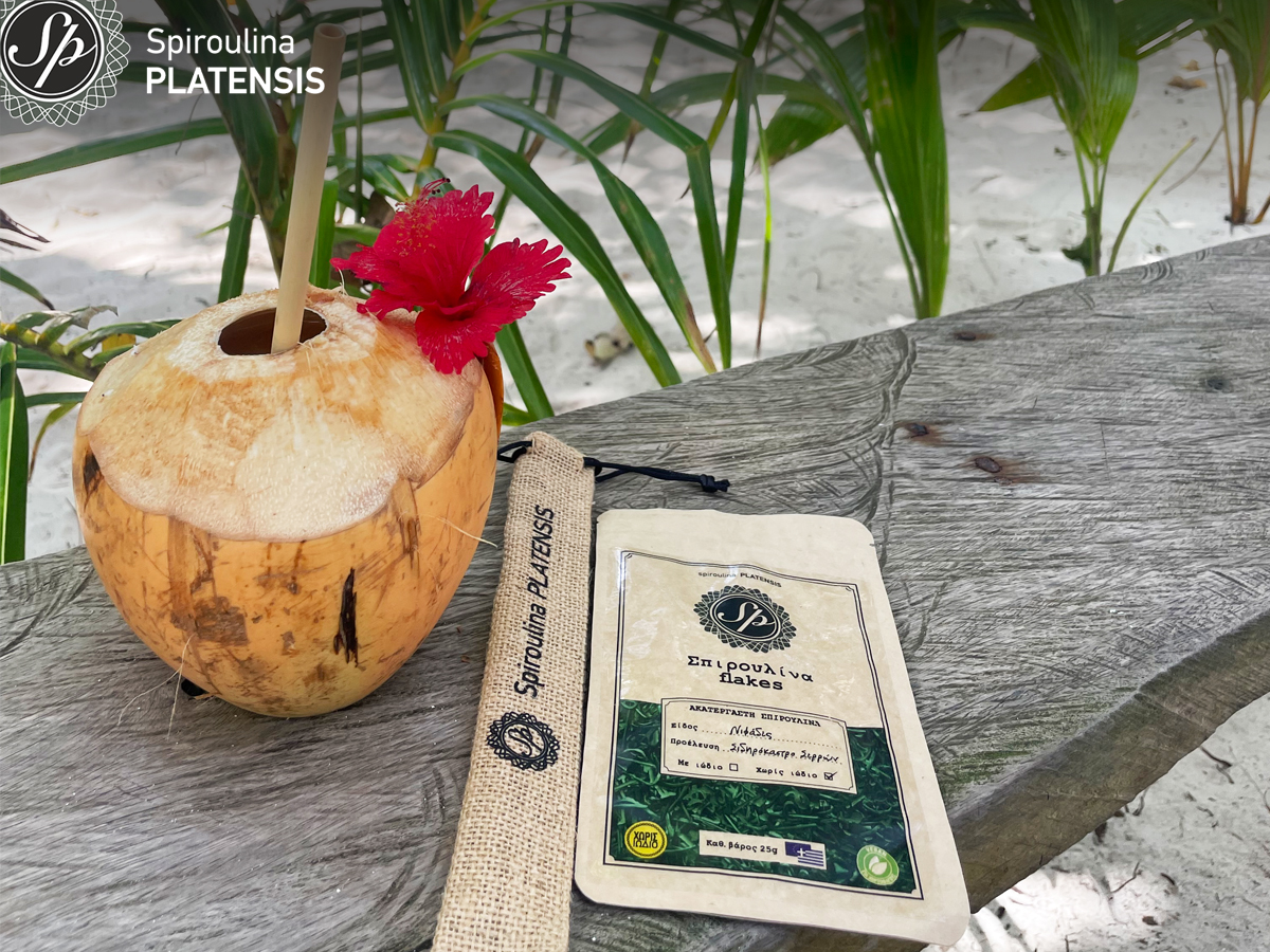 smoothie σε καρύδα πάνω σε ξύλινο πάγκο δίπλα σε ένα bamboo καλαμάκι και μία συσκευασία Σπιρουλίνα flakes