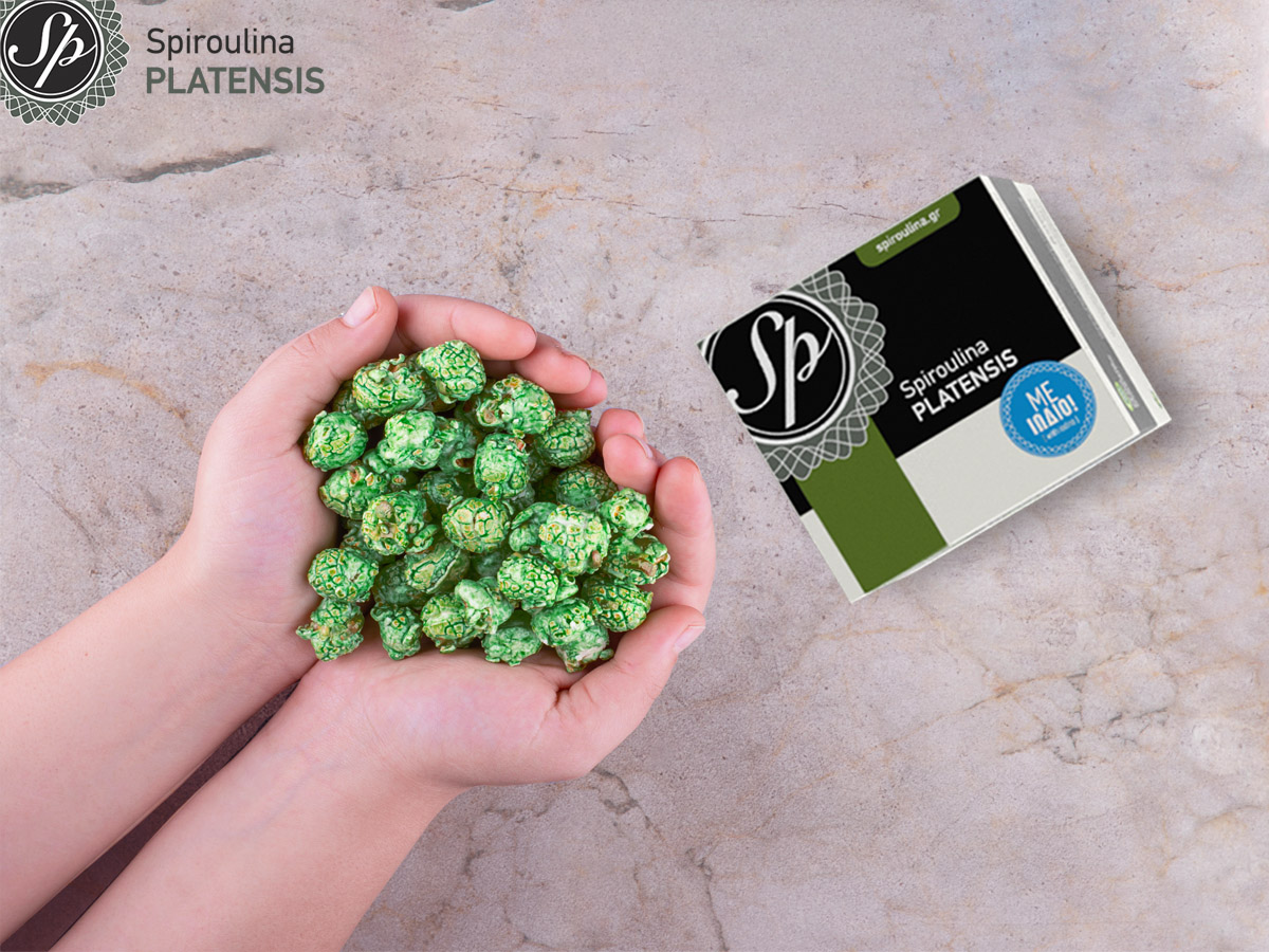 Δύο παιδικά χέρια που κρατούν ποπ κορν σε πράσινο χρώμα σε γκρίζο πάγκο και μία συσκευασία Spiroulina PLATENSIS Σε σκόνη