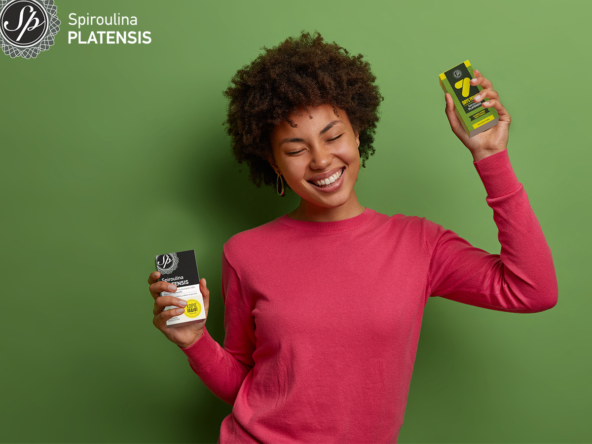 Νεαρή σκουρόχρωμη γυναίκα με ροζ πουλόβερ σε πράσινο φόντο που κρατάει Spiroulina PLATENSIS Ταμπλέτες χωρίς ιώδιο & 7 Days power