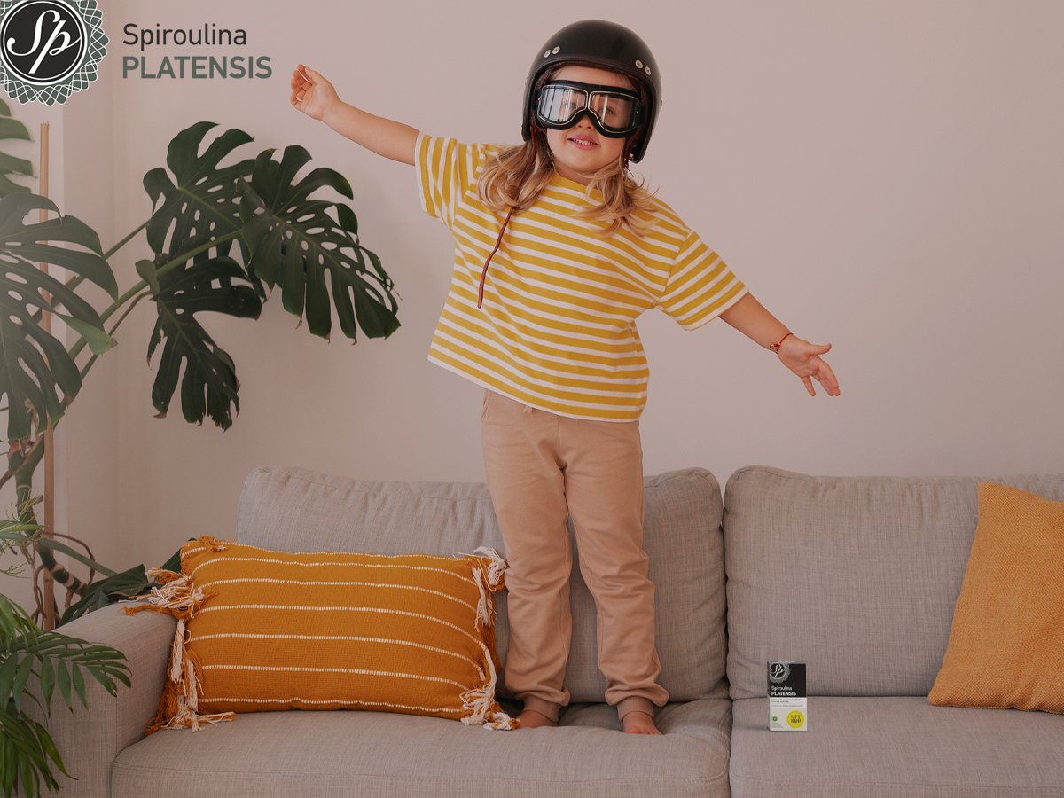 Παιδί με κράνος και γυαλιά παίζει πάνω στον καναπέ, με συσκευασία Spiroulina PLATENSIS δίπλα του στο σαλόνι