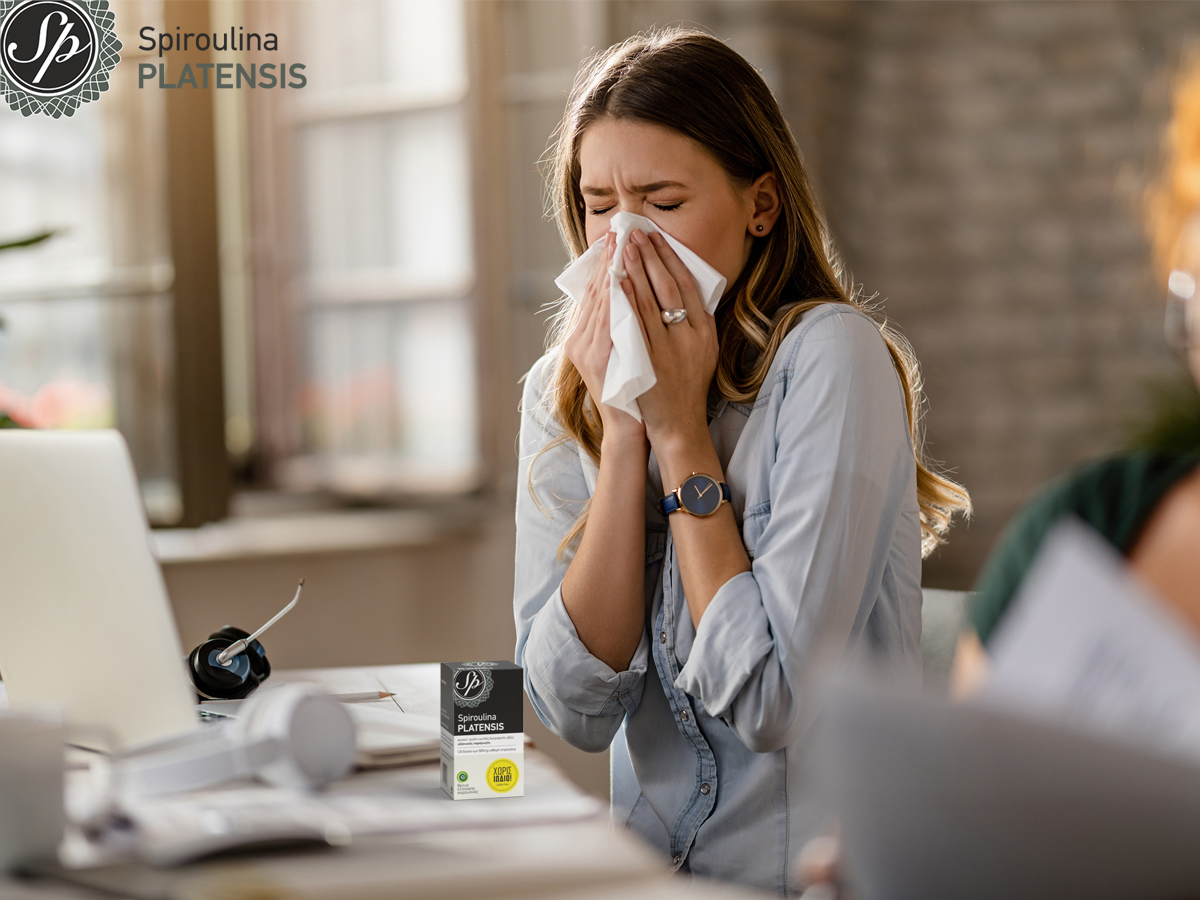 Νεαρή γυναίκα στο γραφείο που φυσάει τη μύτη της επειδή έχει αλλεργία & πάνω στο γραφείο έχει μία συσκευασία Spiroulina PLATENSIS