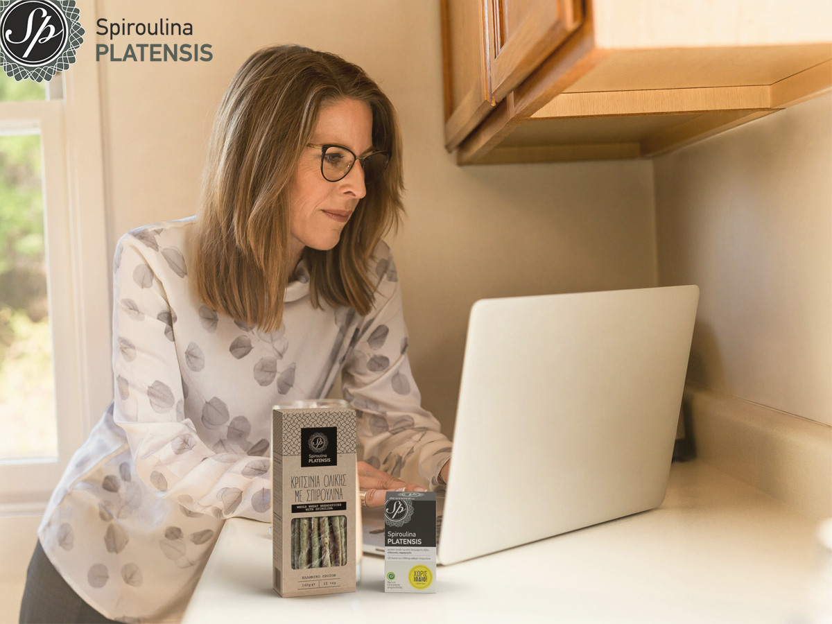 ΄Ώριμη γυναίκα μπροστά στον υπολογιστή της και δίπλα της μία συσκευασία spiroulina PLATENSIS ταμπλέτες & κριτσίνια ολικής με Siproulina PLATENSIS