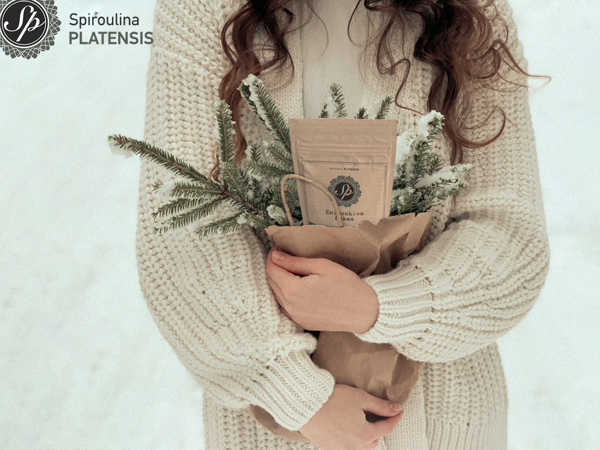 Μία κοπέλα με μπεζ πουλόβερ στα χιόνια που κρατάει μία καφέ χάρτινη σακούλα με κλαριά από έλατο & μία συσκευασία Σπιρουλίνα flakes