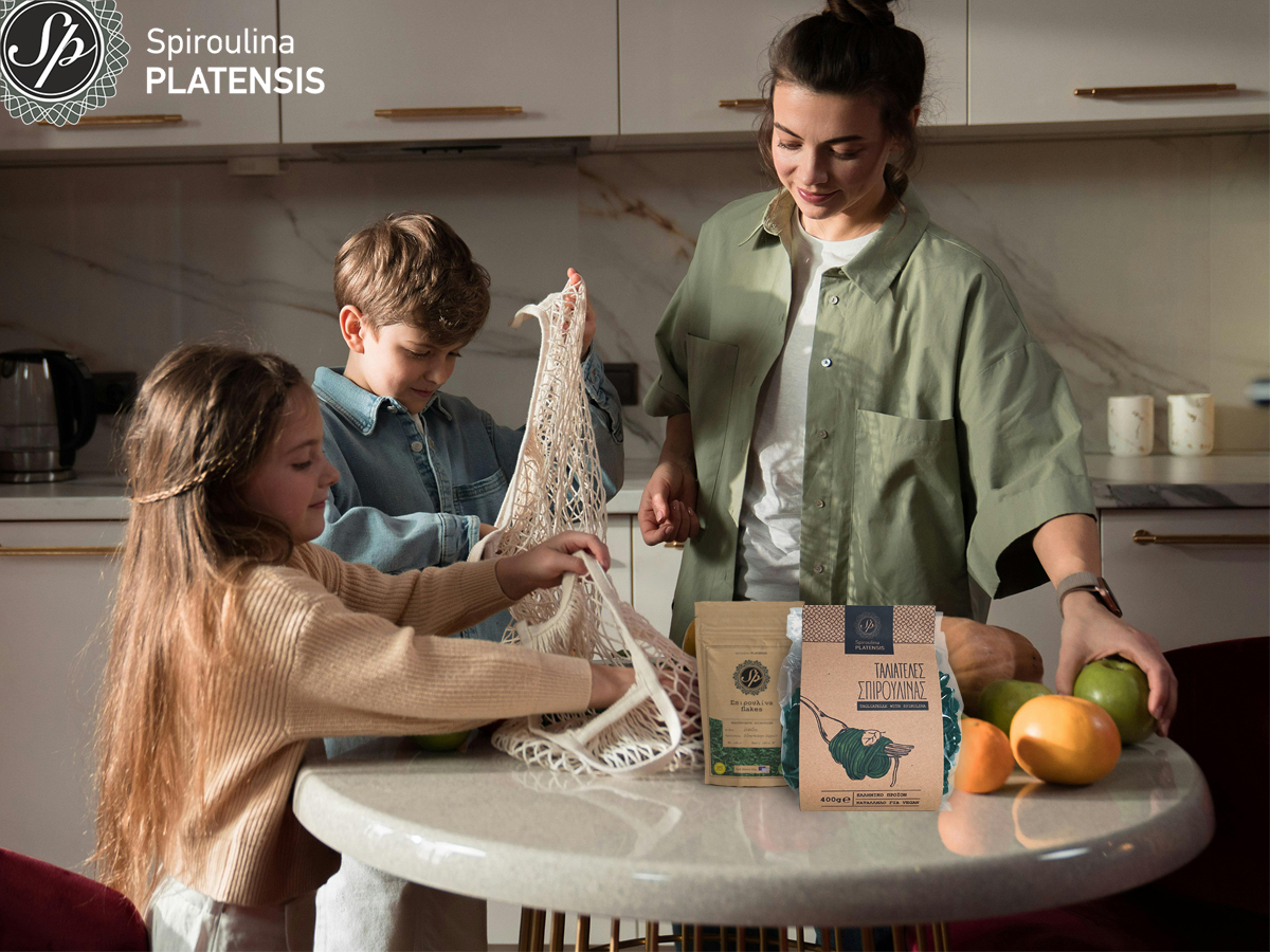 Μία μαμά με τα δύο παιδιά στο τραπέζι με τα ψώνια και πάνω υπάρχουν flakes & ταλιατέλες με spiroulina PLATENSIS