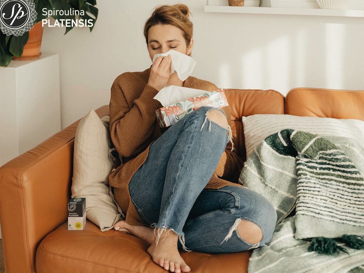 Νεαρή γυναίκα στον καναπέ με καταρροή και μία συσκευασία Spiroulina PLATENSIS ταμπλέτες μπροστά της