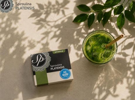Ποτήρι με πράσινο smoothie και συσκευασία Spiroulina PLATENSIS με ιώδιο πάνω σε ανοιχτόχρωμη επιφάνεια με σκιές φύλλων