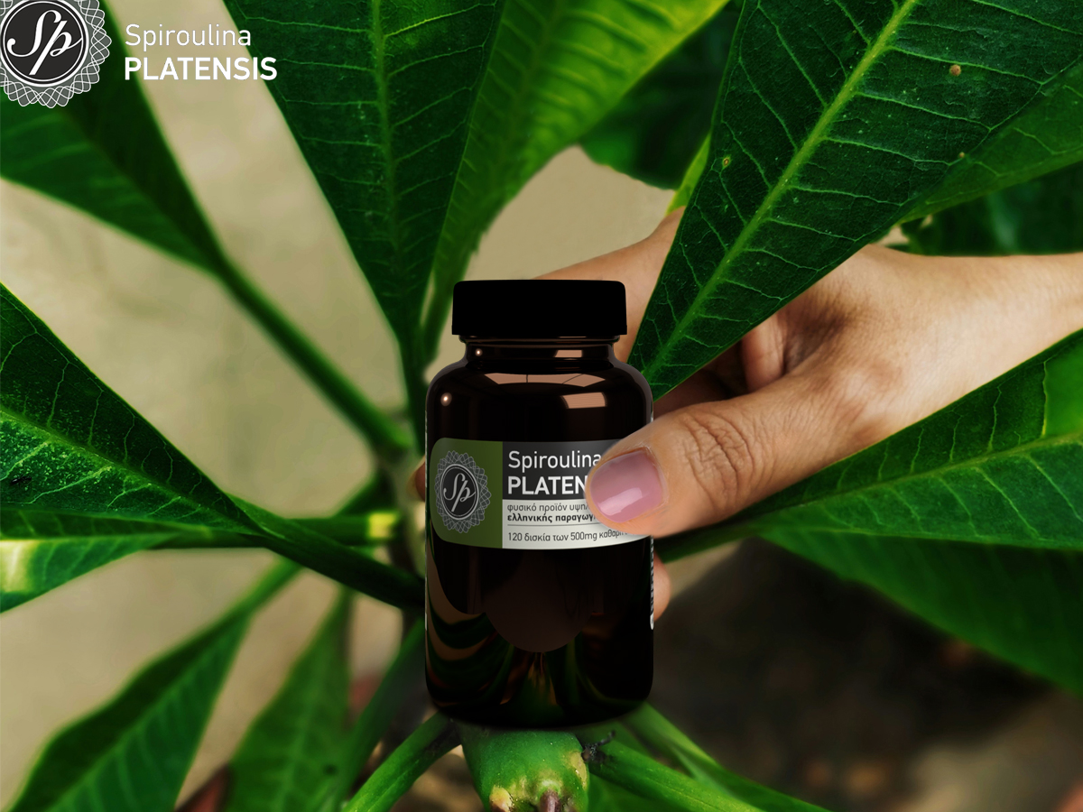 ένα χέρι ανάμεσα σε φύλλα που κρατάει μία συσκευασία spiroulina PLATENSIS ταμπλέτες χωρίς ιώδιο σε γυάλινη συσκευασία